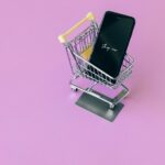 Mobile phone in mini cart on pink surface, symbolizing online shopping.